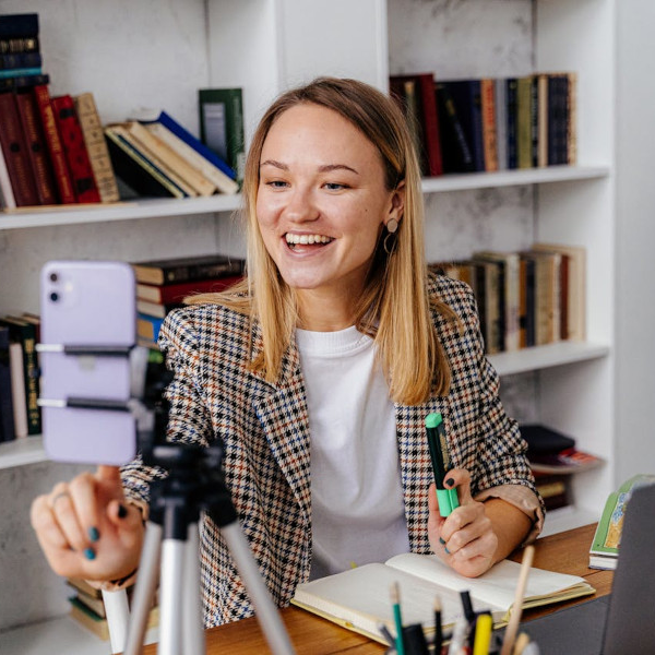 imagen de una mujer sonriendo a la camara de su teléfono móvil mientras maneja varios aparatos tecnológicos más, como un portátil